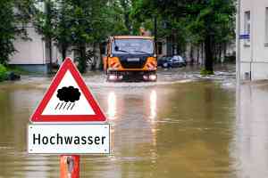 Hochwasserschild vor überfluteter Straße, Straßenmeisterfahrzeug im Hintergrund